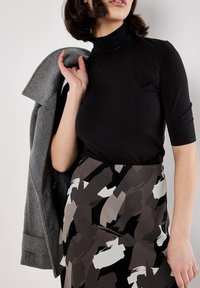 Femme portant un col roulé noir et une jupe à motif abstrait, tenant un manteau gris sur son épaule devant un fond uni.