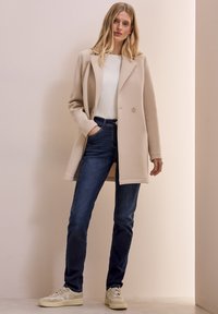 Beige lång kappa med en enda knapp, tillsammans med en vit stickad tröja och mörkblå jeans. Skorna är ljusa sneakers.