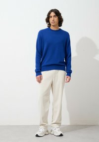 Blauwe gebreide trui met een ronde hals, gecombineerd met lichtbeige ribbroek. Model draagt witte sneakers en staat tegen een neutrale achtergrond.