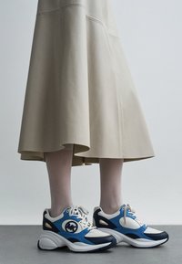 Kék-fehér sportcipők lekerekített orral, hálós betétekkel és mintás talppal. Bézs, térd alatti szoknyával párosítva.