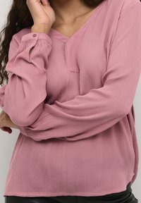 Blouse à col en V en tissu rose doux et texturé avec manches longues dotées de poignets boutonnés et un détail subtil de poche avant.