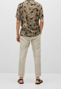 Chemise à manches courtes avec un motif floral marron, associée à un pantalon beige clair retroussé et des sandales marron. Tissu texturé et coupe décontractée.