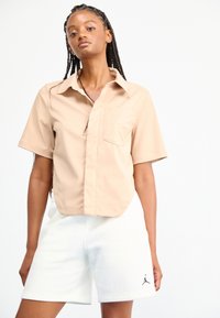 Tan t-shirt met korte mouwen en een voorzak, gecombineerd met witte shorts met een zwart Jordan-logo. Glad materiaal, boxy pasvorm.