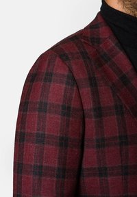 Czerwono-czarna marynarka w kratę z teksturowanej tkaniny, z ostrym kołnierzem i wyraźnie zarysowanymi ramionami, cięta pod kątem.