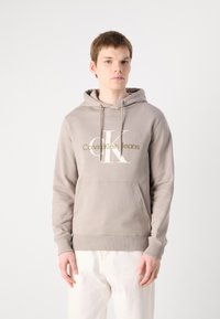 Felpa color beige chiaro realizzata in tessuto morbido, con una tasca a kangaroo sul davanti e un logo "Calvin Klein Jeans" in bianco e oro.