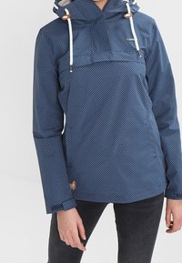 Granatowa wodoodporna kurtka w białe kropki, z regulowanym kapturem, sznurkiem i dwukolorowymi akcentami. Miękka, gładka tekstura z luźnym krojem.