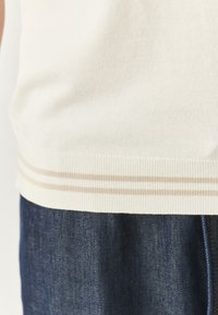 Maglione lavorato a maglia crema con orlo a coste, caratterizzato da strisce beige; abbinato a jeans in denim blu scuro, che mostrano contrasto di texture e colore.