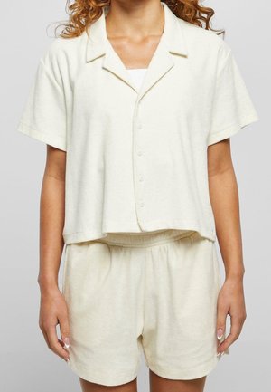 Woman wearing a cream short-sleeve button-up shirt with collar and matching elastic-waist shorts against a gray background.