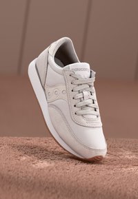 Světlé šedé tenisky z mesh a semiše, s texturovaným svrškem, šněrováním a bílou gumovou podrážkou s hnědými akcenty.