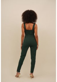 Kvinna med lockigt hår står vänd bortåt, klädd i en mörkgrön figurnära jumpsuit och beige högklackade sandaler mot en enfärgad bakgrund.