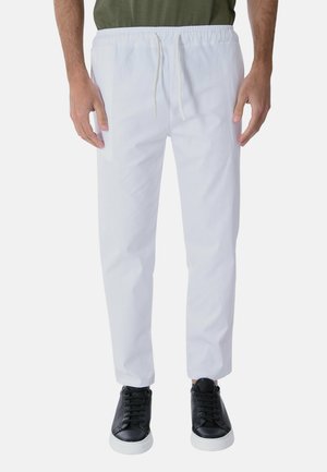 Uomo con pantaloni bianchi con coulisse, sneakers nere e camicia verde oliva, in piedi davanti a uno sfondo chiaro e uniforme.