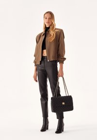 Giacca bomber marrone con polsini elastici, top corto nero, pantaloni di pelle nera e borsa nera trapuntata con manici a catena.
