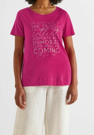 Fuksianpunainen lyhythihainen t-paita, jossa on pyöreä pääntie ja metallinen tekstikuvio. Yhdistetty vaaleanbeigeihin pellavahousuihin.