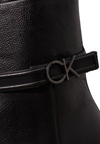 Gros plan d'une botte en cuir noir texturé avec une fine sangle et une boucle argentée avec le logo « CK » sur le côté.