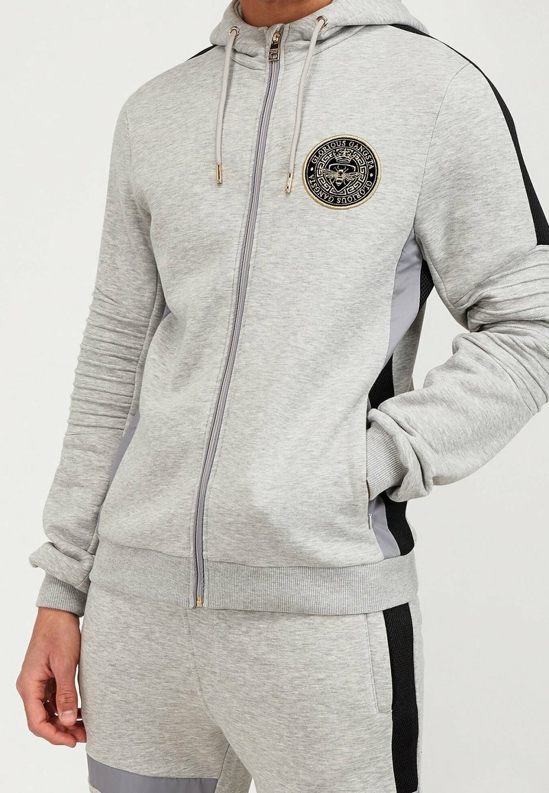 Homme portant un sweat à capuche gris clair zippé et un jogging assorti avec des panneaux latéraux noirs et gris et un emblème rond sur la poitrine.