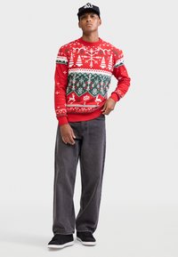 Maglione rosso con motivo a maglia con fiocchi di neve bianchi, alberi verdi e renne; abbinato a jeans grigi e scarpe da ginnastica nere.