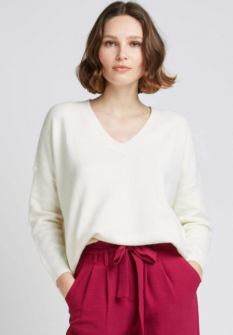 Pull en V blanc avec une texture douce et une coupe ample, associé à un pantalon rose taille haute avec un nœud à l'avant et des poches.