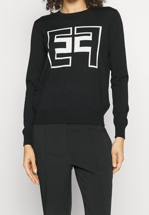 Person iført en sort langærmet sweater med hvidt geometrisk "FF" logo-design og sorte bukser, der står mod en ensfarvet baggrund.