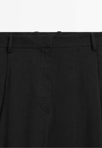 Pantalon noir formel avec passants de ceinture et fermeture éclair frontale, montrant la partie haute de la taille.