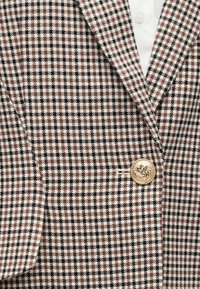 Blazer pied-de-poule marron, noir et blanc avec un bouton doré gravé d’un motif de couronne, porté sur une chemise blanche.