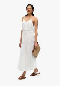 Hvid, ærmeløs maxikjole med blonder detaljer, tiered kant og et let design. Båret med brune sandaler og holder en vævet clutch.