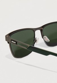 Gafas de sol de la marca Hugo con lentes de color verde oscuro, montura metálica negra mate y brazos de plástico verde con detalle del logo en el lateral.