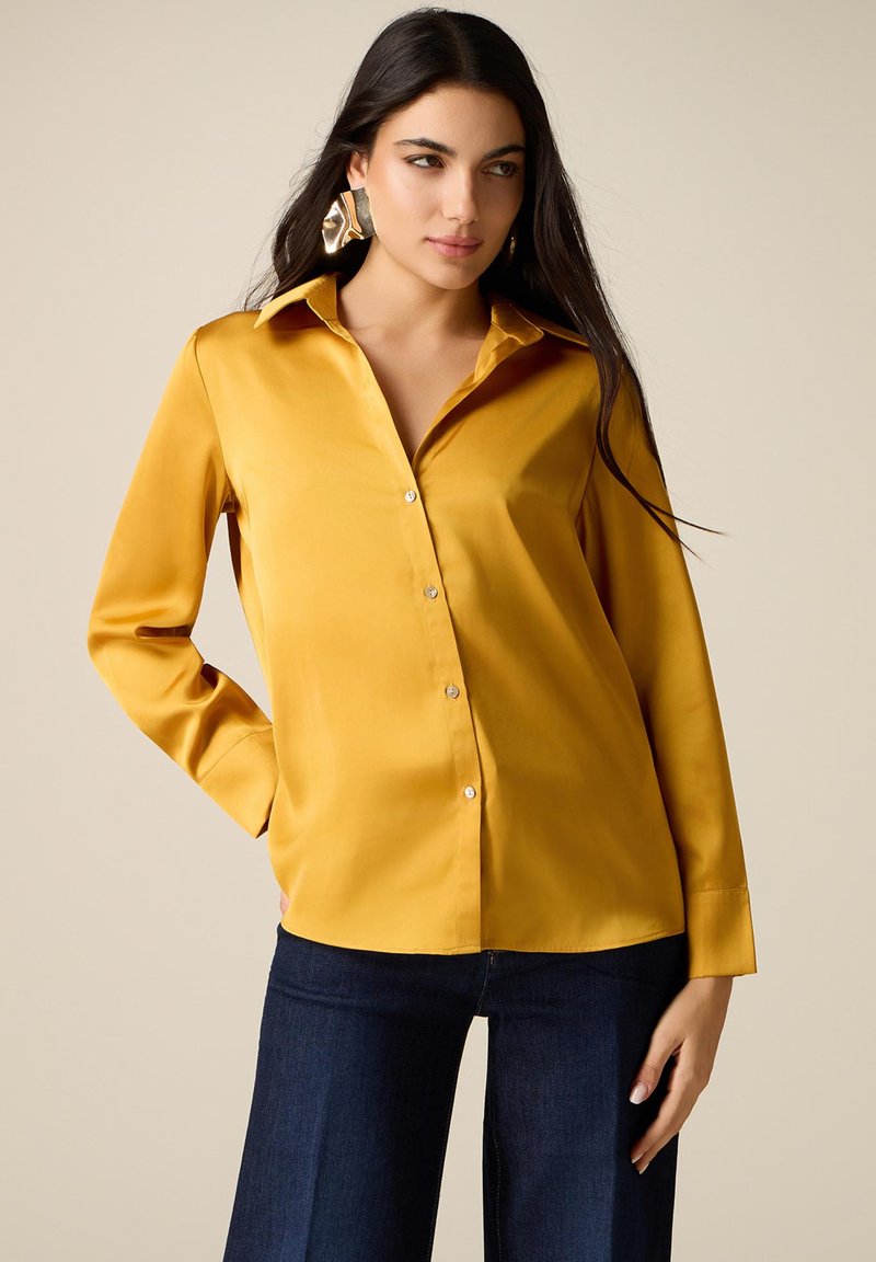 Mujer con cabello largo y oscuro, vistiendo una camisa satén color mostaza con botones, jeans oscuros y grandes pendientes abstractos dorados, de pie con una mano en el bolsillo.