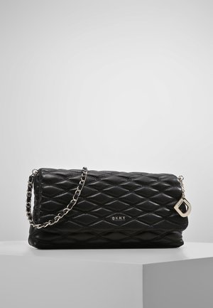 Zwarte gewatteerde leren DKNY-handtas met een zilveren kettingriem en een metalen D-bedel, geplaatst op een wit oppervlak tegen een lichtgrijze achtergrond.