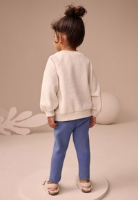 Bluză crem cu textură și mâneci bufante, asortată cu pantaloni sport albastre și încălțăminte deschisă la culoare. Ținută simplă și casual, privită din spate.