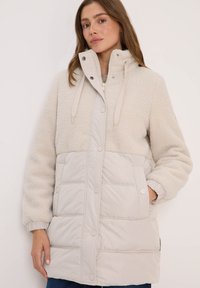 Veste légère beige avec un haut en polaire et une partie inférieure rembourrée. Comprend une capuche, des boutons-pression et des poches latérales.