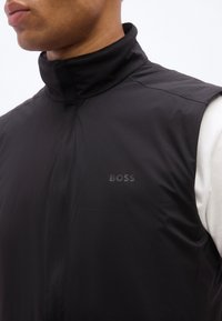 Άνδρας που φοράει μαύρο αμά νικο γιλέκο με ψηλό γιακά και το λογότυπο "BOSS" στο στήθος πάνω από ένα λευκό μακρυμάνικο πουκάμισο.