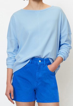 Persoon draagt een lichtblauw shirt met lange mouwen, gedeeltelijk ingestopt in felblauwe, hooggesloten korte broek, met één hand in de zak.