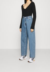 Mustavärinen ribattu pitkähihainen toppi V-aukkolla, yhdistettynä korkeavyötäröisiin vaaleansinisii leveisiin farkkuihin ja valkoisiin lenkkareihin, joissa on mustia yksityiskohtia.