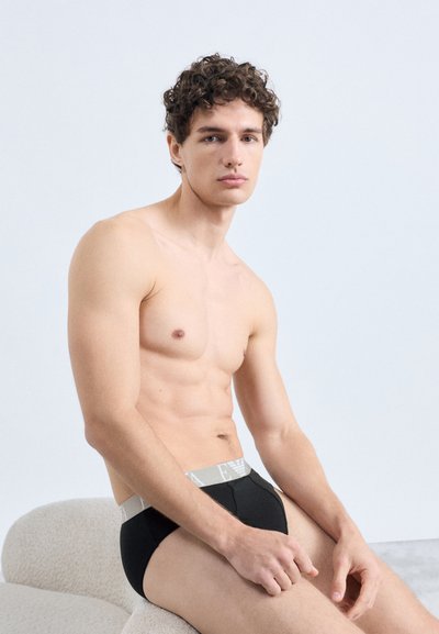 Fekete alsónadrág szürke derékrésszel, amely logós feliratot tartalmaz. Modell rövid göndör hajjal és meztelen felsőtesttel, egy texturált világos háttér előtt ül.