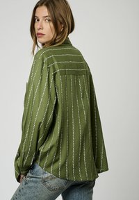 Camicia verde oliva con strisce verticali bianche, caratterizzata da una vestibilità ampia, maniche lunghe e un dettaglio di yoke sul retro. Tessuto morbido.