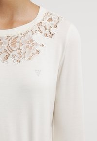 Vit långärmad topp med en spetsinlägg i blommönster, slät textur, rund halsringning och liten triangulär logotypdetalj.