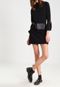 Abito nero a maniche lunghe con dettagli a taglio, abbinato a una borsa a cintura nera e stivali alla caviglia lucidi. Tessuto morbido, forma aderente.