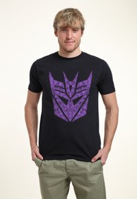 Zwarte T-shirt met een groot paars Transformers-logo op de voorkant. Korte mouwen, ronde hals, gemaakt van katoen. Casual pasvorm.