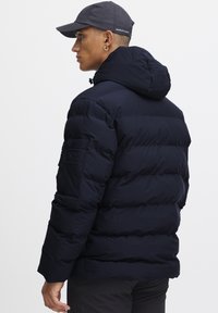 Mørkeblå puffjakke med hætte, der har en lynlås på ærmet og en quiltet design, lavet af et blødt, struktureret materiale.