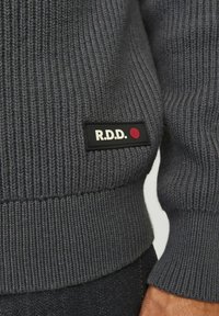 Hallux hallitus riidepuudega elastset kampsunit, millel on must silt "R.D.D." ja punane punkt, mis on esitatud vasakus alanurgas.