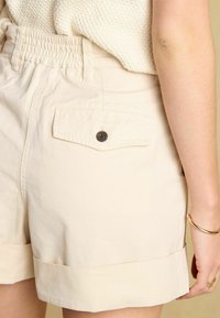 Beżowe bawełniane szorty z elastycznym pasem, podwiniętymi nogawkami oraz tylną kieszenią z czarnym guzikiem. Gładka faktura.