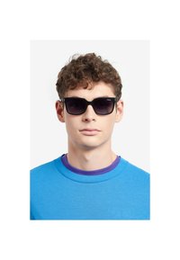 Ochelari de soare negri cu lentile întunecate, cadru rectangular și mici accente aurii. Purtați de o persoană în hanorac albastru cu guler violet.