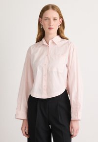 Camicia button-up corta di colore rosa chiaro, con maniche lunghe, colletto e taschino sul petto. Realizzata in un tessuto liscio con una leggera lucentezza.
