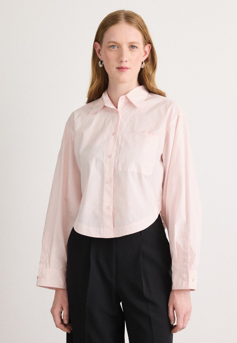 Camicia button-up corta di colore rosa chiaro, con maniche lunghe, colletto e taschino sul petto. Realizzata in un tessuto liscio con una leggera lucentezza.