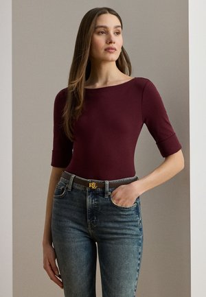 Kvinde med langt brunt hår iført en bordeaux tætsiddende top med albuelange ærmer og højttaljede blå jeans med sort bælte.
