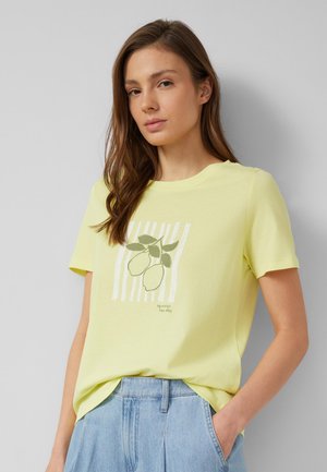 Jonge vrouw met lang bruin haar, draagt een lichtgeel T-shirt met citroen- en bladprint en lichtblauwe spijkerbroek.