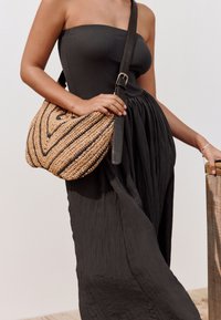 Borsa intrecciata in paglia con strisce nere, forma curva e tracolla regolabile in pelle, tenuta accanto a un abito nero fluente.