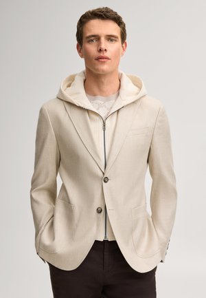 Jonge man draagt een beige blazer over een lichte hoodie met rits en een donkere broek, staand met handen in de zakken tegen een effen achtergrond.