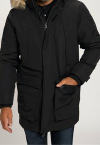Parka noire avec capuche bordée de fourrure. Comprend une fermeture éclair et des boutons, plusieurs poches avant, et des poignets côtelés. Tissu texturé.