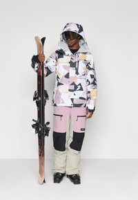 Suusakomplekt, mis sisaldab mitmevärvilist mustriga jopet kapuutsiga, koos roosa ja musta värvi pükstega; inimene hoiab suusavarustust.